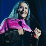 a woman with long gray hair and pink scarf standing in front of a microphone