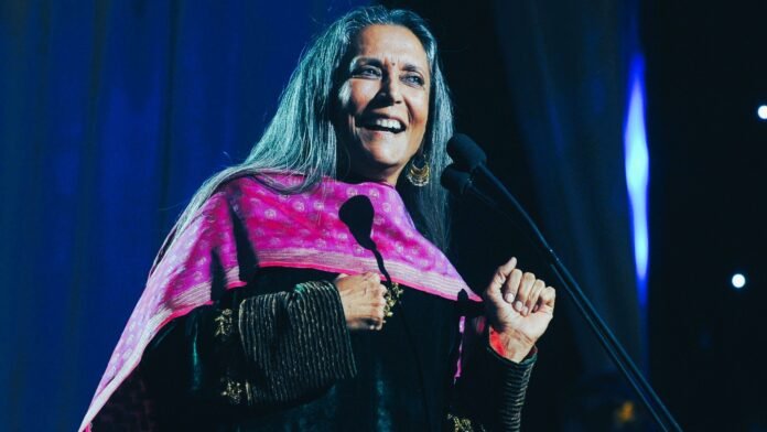 a woman with long gray hair and pink scarf standing in front of a microphone